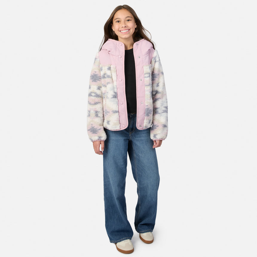 A young girl stands against a light gray studio background wearing a fuzzy hooded jacket with a pink hood and collar, a pastel tie-dye pattern on the sleeves and body, a black top, wide-leg blue jeans, and white slip-on shoes.