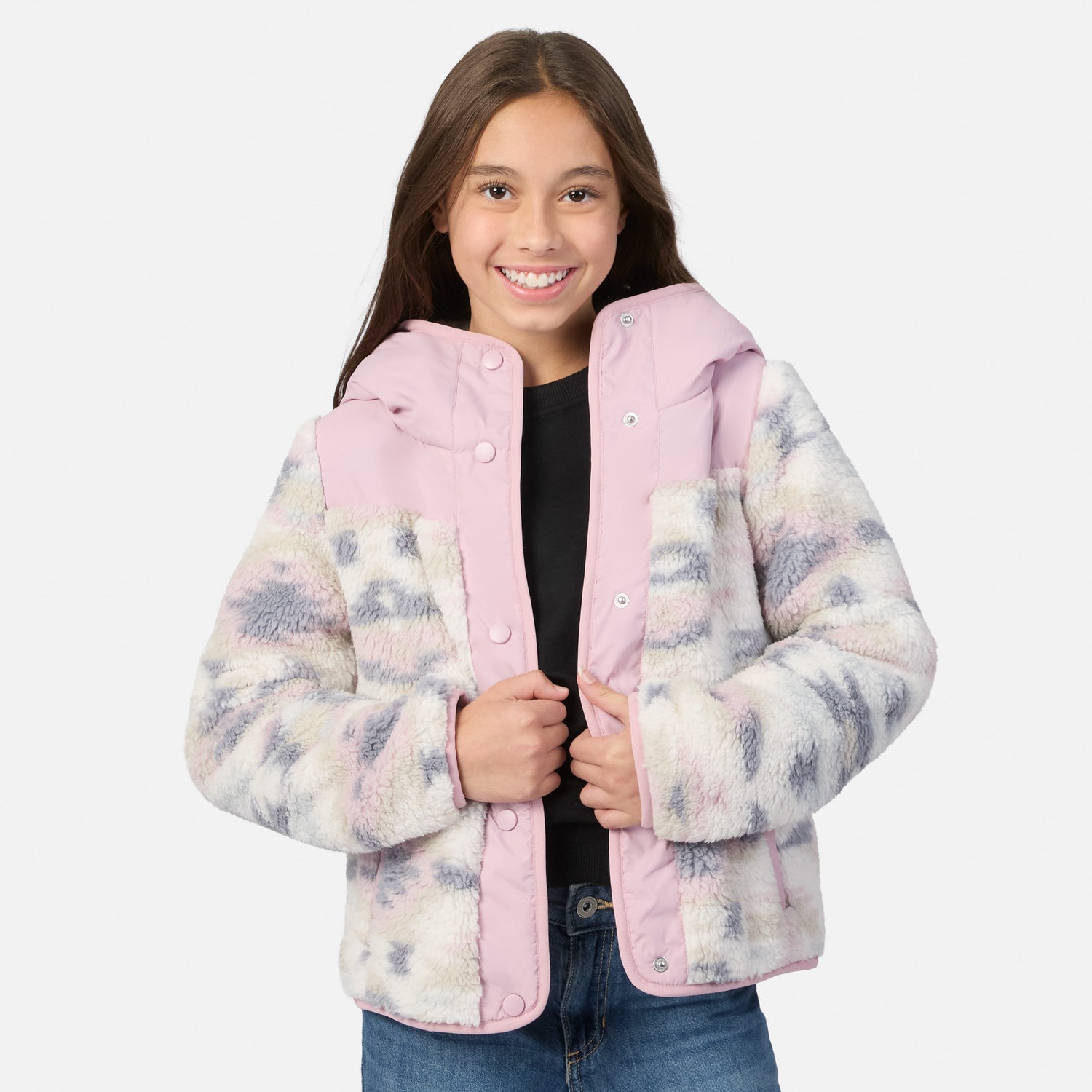 A young girl smiling while wearing a pink and white faux fur jacket with a pastel tie-dye pattern on the sleeves, paired with a black top and blue jeans, against a light gray background.