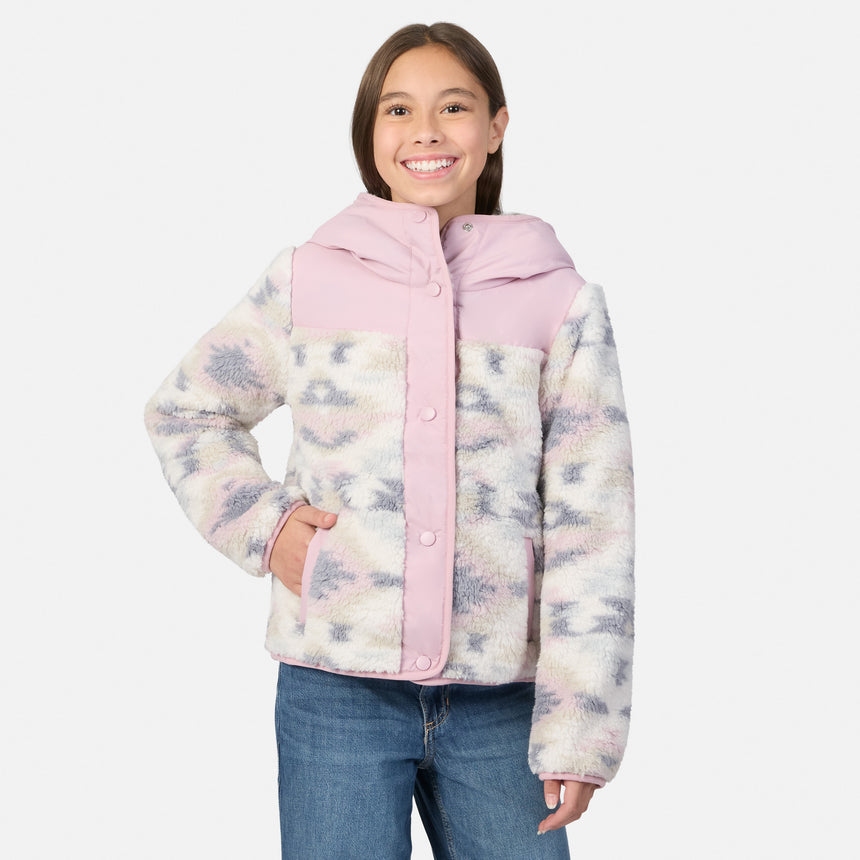 A young girl smiling while wearing a pink and patterned sherpa fleece jacket with blue jeans, photographed against a light gray studio background.