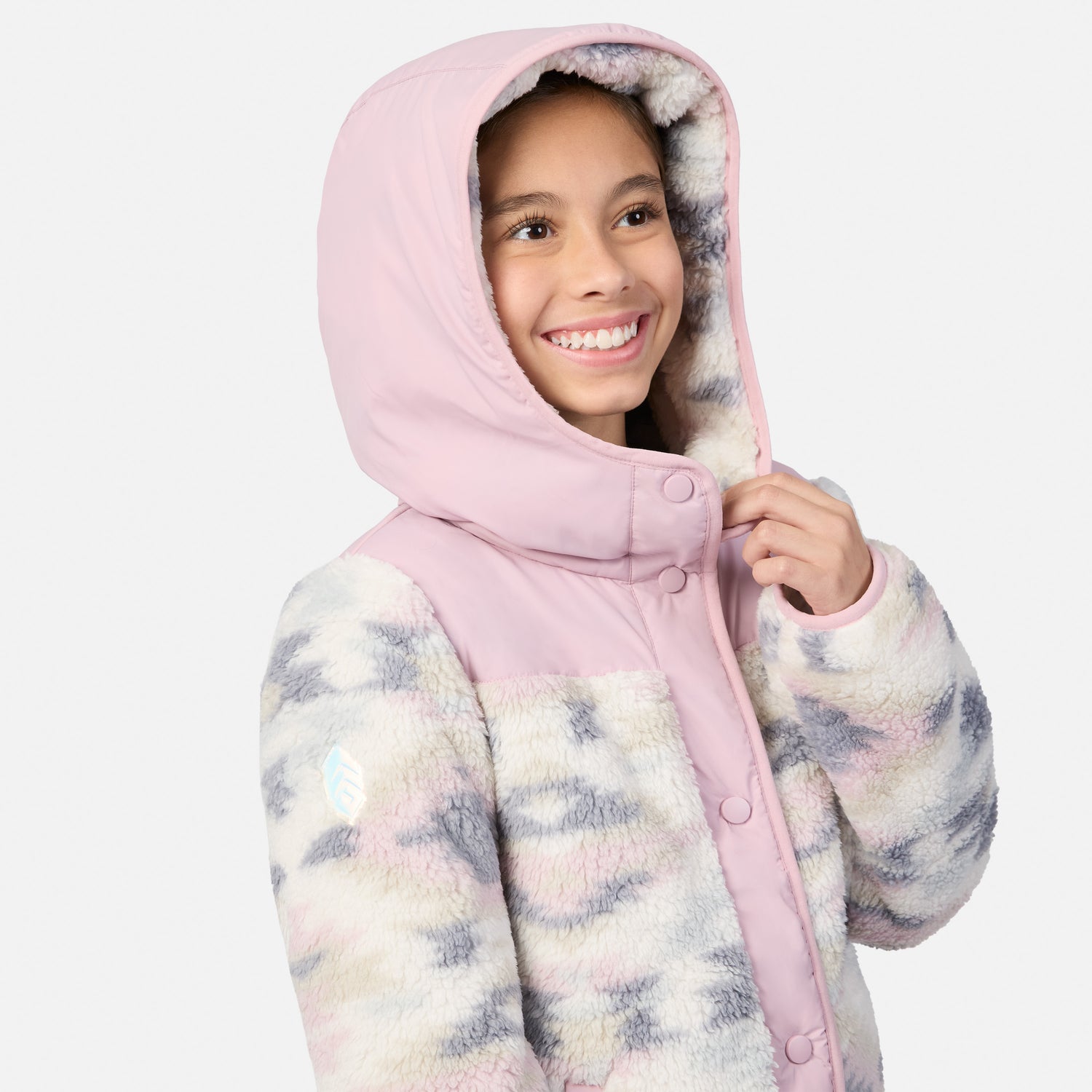 A smiling girl wearing a hooded jacket with a pink hood and collar, and a fuzzy pastel tie-dye body in pink, blue, and cream, photographed against a light gray studio background.