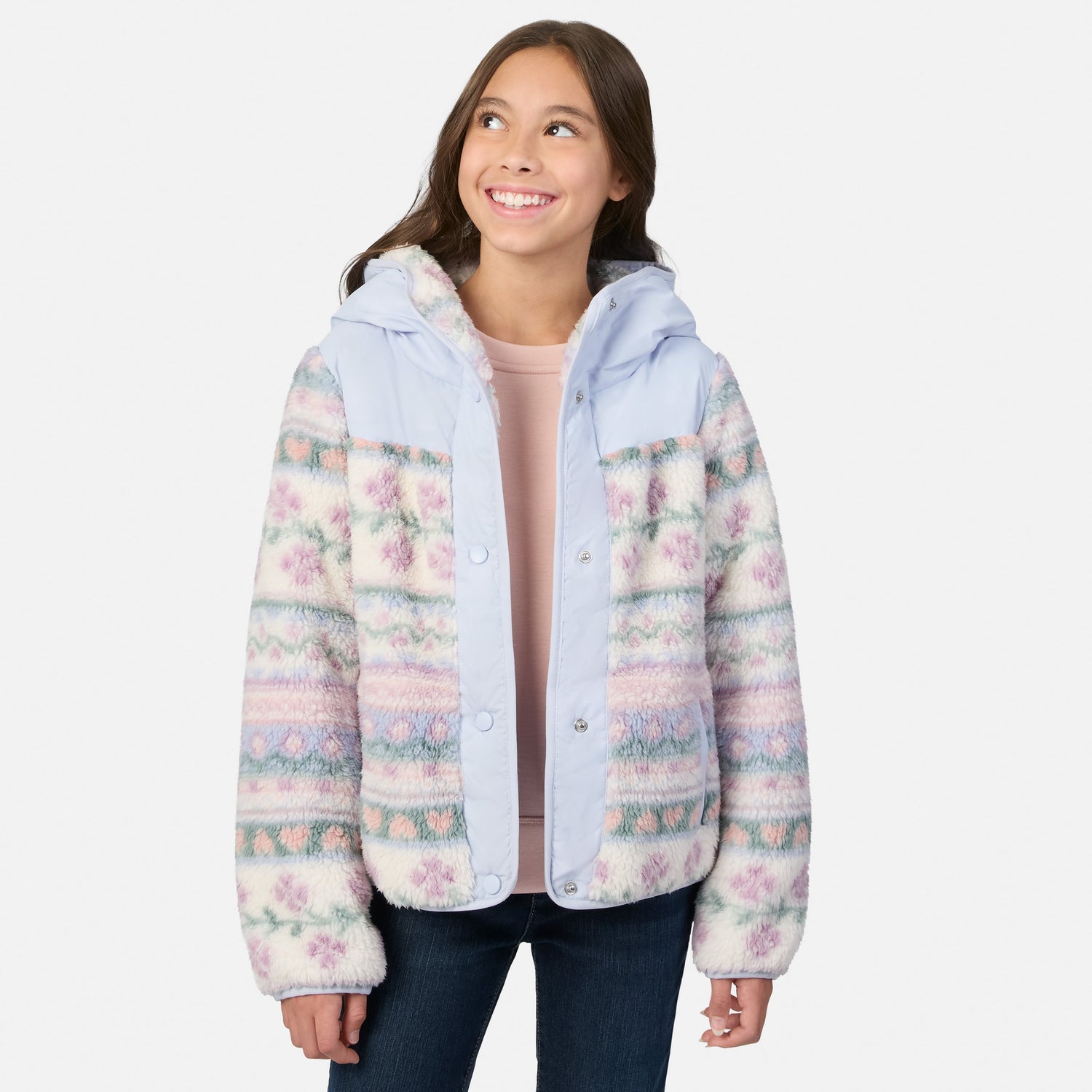 A young girl smiling and looking up while wearing a light blue hooded jacket with a pastel multicolor Fair Isle pattern on the sleeves and body, paired with a pink top and dark blue jeans, against a light gray background.