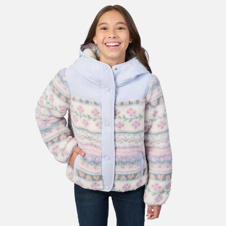 A smiling young girl wearing a hooded fleece jacket with a pastel multicolor floral pattern and light blue trim, paired with dark blue jeans, against a plain light gray background.