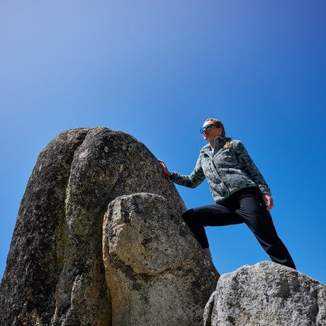 A woman wearing a gray patterned fleece jacket and black pants climbs large rock formations under a clear blue sky.