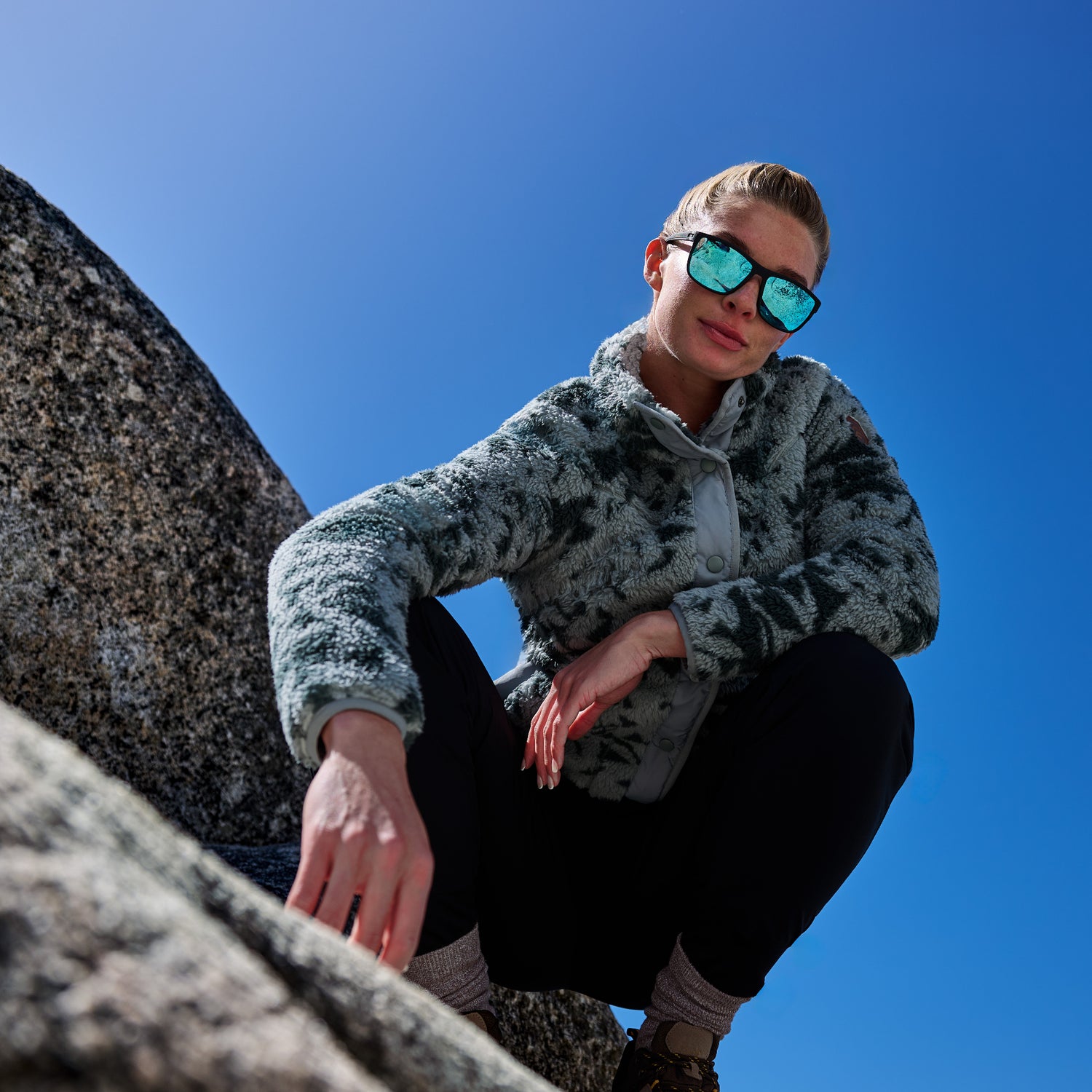 A woman crouching outdoors on a rock against a clear blue sky, wearing a fuzzy gray and black patterned fleece jacket, black pants, and mirrored sunglasses.