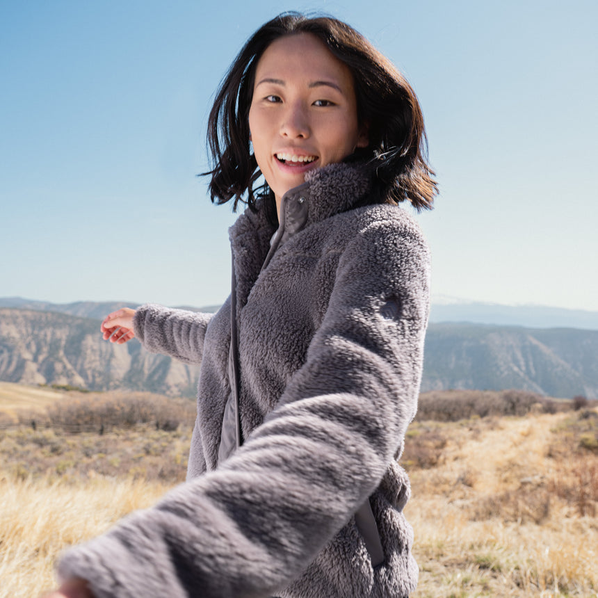 Woman in a gray fleece jacket standing in a desert landscape with mountains in the background.