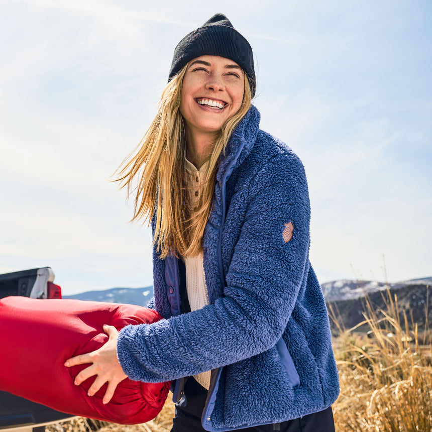 Person wearing a blue jacket and black beanie holding a red object outdoors with mountains in the background