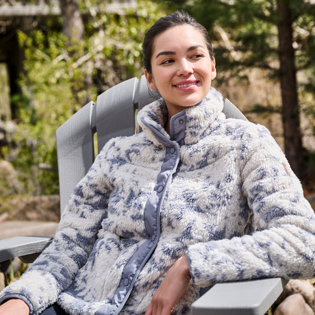 A woman wearing a textured, high-collar fleece jacket in a blue and white patterned design, sitting in a gray outdoor patio chair with a blurred forest background.