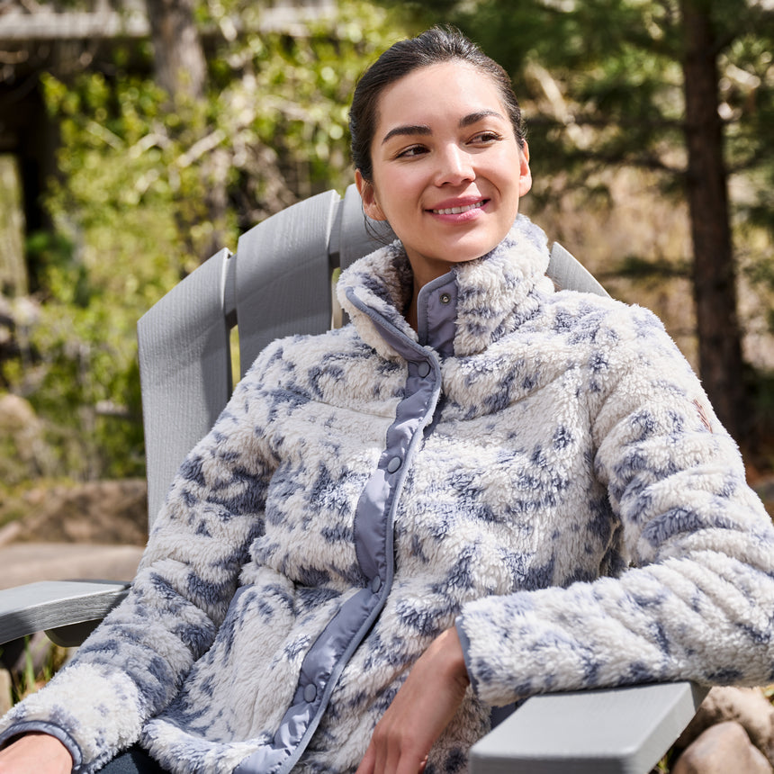 A woman wearing a textured, high-collar fleece jacket in a blue and white patterned design, sitting in a gray outdoor patio chair with a blurred forest background.