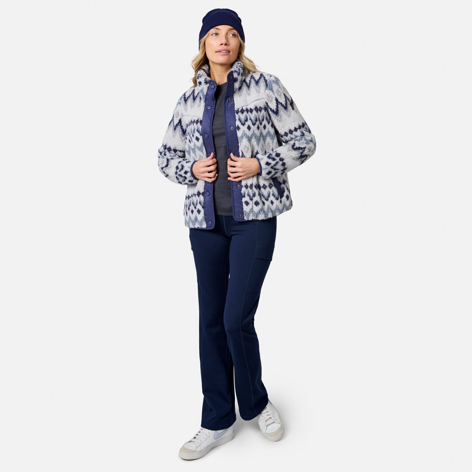 [Model is wearing size S.] A woman modeling a patterned zip-up fleece jacket, navy pants, a navy beanie, and white sneakers against a light gray studio background.