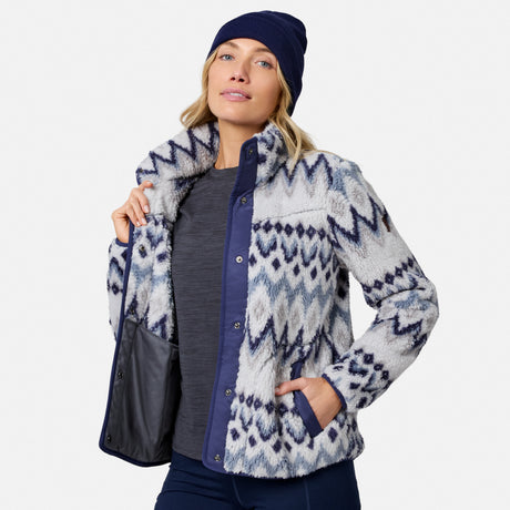 [Model is wearing size S.] A woman wearing a navy blue beanie, a gray long-sleeve top, and a fuzzy zip-up jacket with a blue and white geometric pattern. She is holding a black shoulder bag against a light gray studio background.
