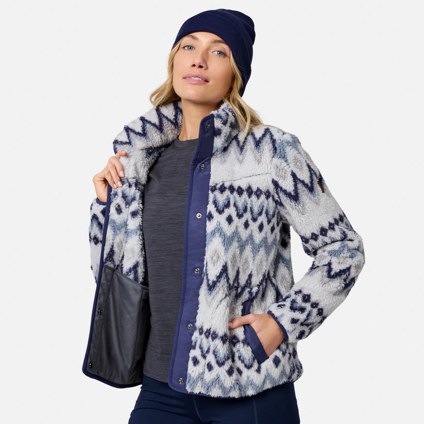 [Model is wearing size S.] A woman wearing a navy blue beanie, a gray long-sleeve top, and a fuzzy zip-up jacket with a blue and white geometric pattern. She is holding a black shoulder bag against a light gray studio background.