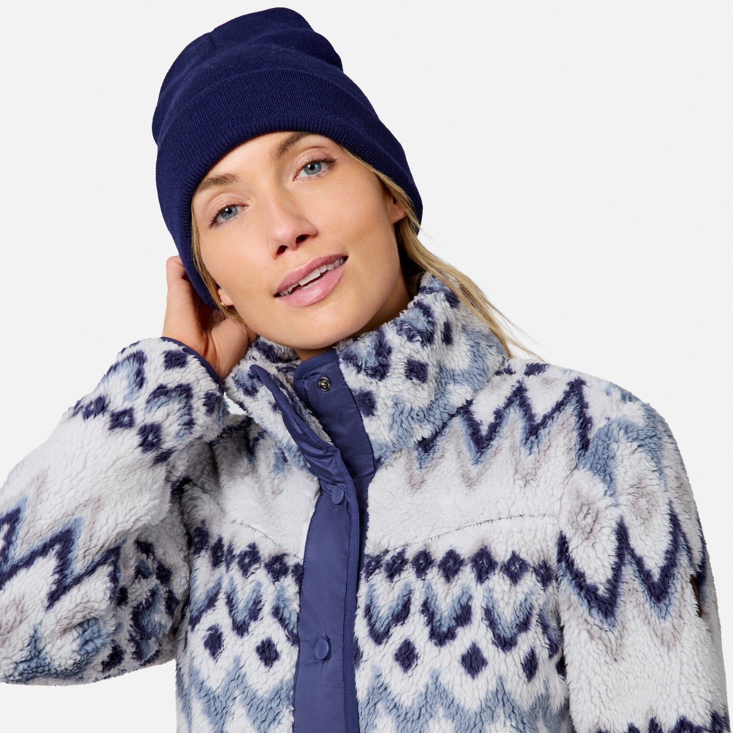 [Model is wearing size S.] A woman wearing a navy blue knit beanie and a fuzzy zip-front fleece jacket with a blue and white geometric pattern, photographed against a light gray studio background.