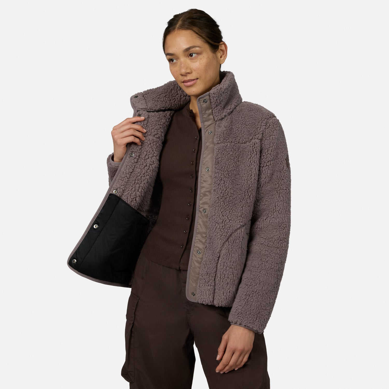 [Model is wearing size S.] A woman modeling a taupe brown sherpa fleece jacket with a high collar and snap-button front, styled over a dark brown top and pants against a light gray studio background.