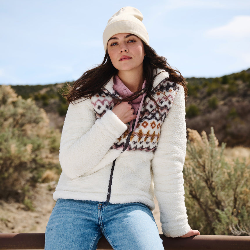 Woman wearing a white jacket with a patterned design, blue jeans, and a pink scarf outdoors.