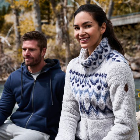 A woman in a Free Country Women’s Sierra Butter Pile® Zip Up Jacket smiles outdoors by a river with a man in a blue hoodie, both surrounded by vibrant autumn trees.