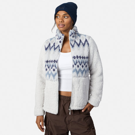 A woman models the Free Country Women’s Sierra Butter Pile® Zip Up Jacket in a white and gray pattern, styled with a white tank, brown pants, and navy beanie, standing against a plain background with one hand in her pocket.