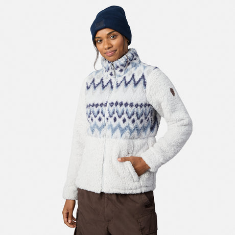 A person wears a Free Country Women’s Sierra Butter Pile® Zip Up Jacket in Alpine Aztec Dusk, with a navy beanie and brown pants, standing with one hand in their pocket against a plain background.