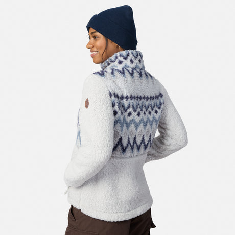 A person in a navy beanie and the Free Country Women’s Sierra Butter Pile® Zip Up Jacket with blue zigzag patterns smiles, facing backwards with one hand on their hip against a plain background.