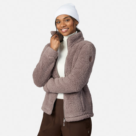 A woman in a white beanie, cream turtleneck, and mauve Women’s Sierra Butter Pile® Zip Up Jacket by Free Country smiles with arms crossed, standing against a plain white background.