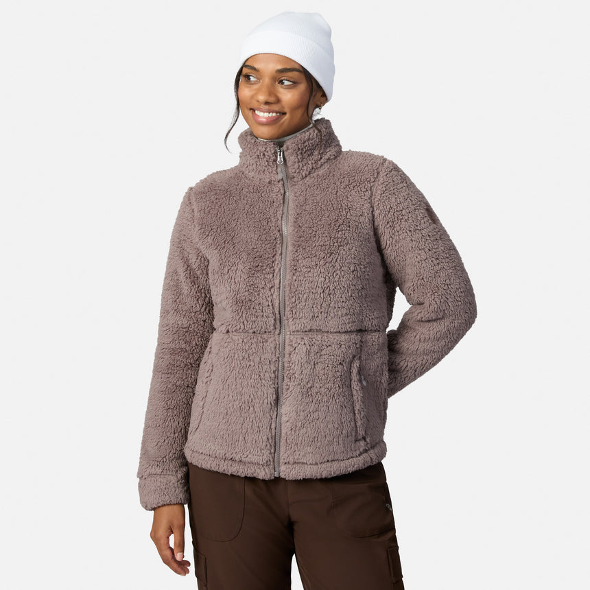 A woman smiles with one hand on her hip wearing brown pants and a Women’s Sierra Butter Pile® Zip Up Jacket by Free Country in Zinc, styled with a white beanie against a light background.