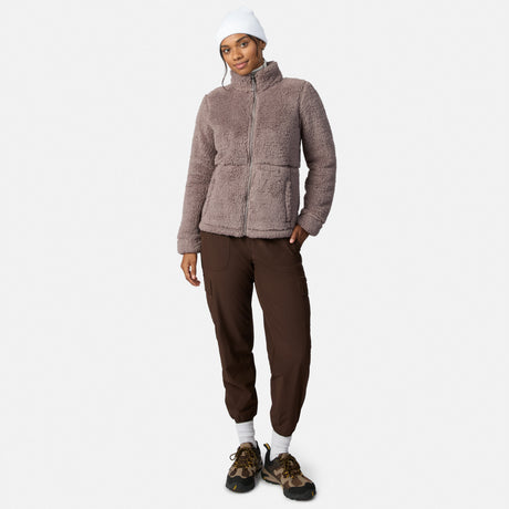 A person models the Free Country Women’s Sierra Butter Pile® Zip Up Jacket in mauve with a white beanie, dark brown joggers, white socks, and brown hiking shoes against a plain white background.