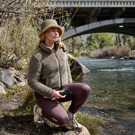 A woman kneels by a river under a concrete bridge, wearing a textured fleece zip jacket, olive green bucket hat, mauve-brown pants, and beige hiking boots while holding a black camera.