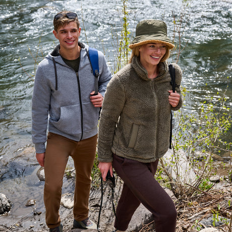 A man and a woman hiking outdoors by a flowing river. The woman wears an olive green sherpa fleece zip-up jacket, brown hiking pants, and an olive green bucket hat.