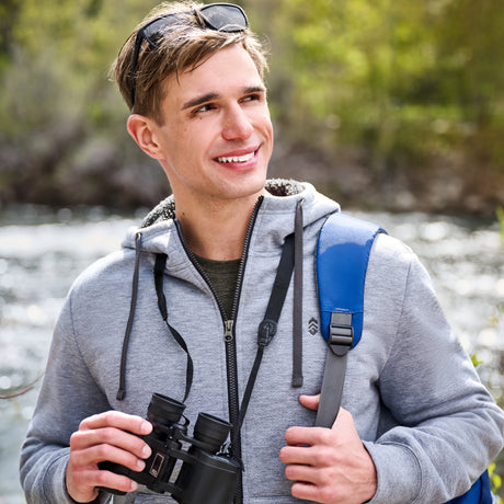 A young man wearing the Free Country Everybody Sherpa Bonded Hoodie in grey stands outdoors near water, smiling and holding binoculars. He has sunglasses resting on his head and carries a backpack with blue straps. The background is filled with softly blurred trees and greenery.