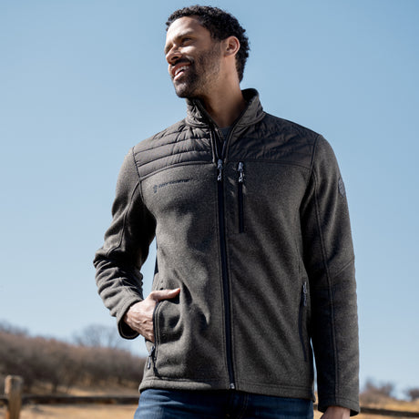 A man enjoys a sunny day outdoors, smiling as he looks aside. He wears the Free Country Men's Frore Knit Fleece Jacket with quilted shoulders and jeans, one hand in his pocket. Sparse trees and blue sky complete the background.