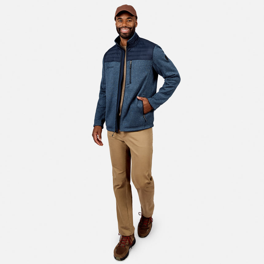 A man in the Free Country Men’s Frore Knit Fleece Jacket, blue with a convertible collar, plus brown pants, shoes, and cap, smiles while walking against a plain white background.