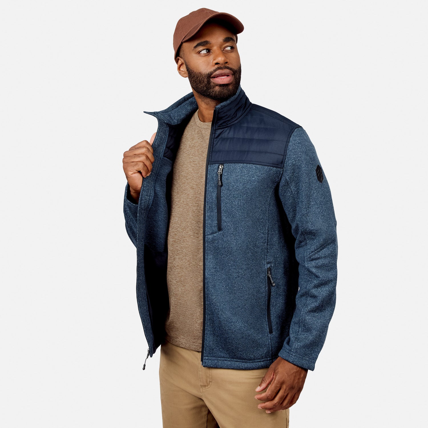 A man wears the Free Country Men’s Frore Knit Fleece Jacket—blue with black shoulder detail and convertible collar—over a beige shirt, tan pants, and a brown cap, standing against a plain white background, looking slightly to the side.