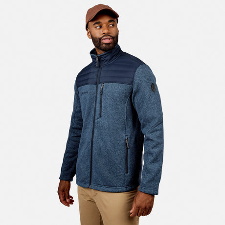 Man wearing a brown cap, beige pants, and the Free Country Men’s Frore Knit Fleece Jacket in Cadet with black shoulder panels and zippered pockets, standing against a plain white background and looking to his left.