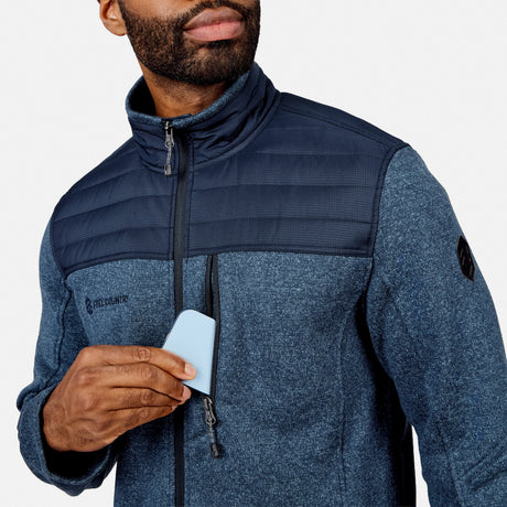 A man in a Free Country Men’s Frore Knit Fleece Jacket with a convertible collar unzips the chest pocket to put in a light blue card. His face, partially shown, has a short beard and mustache. The background is plain and light.