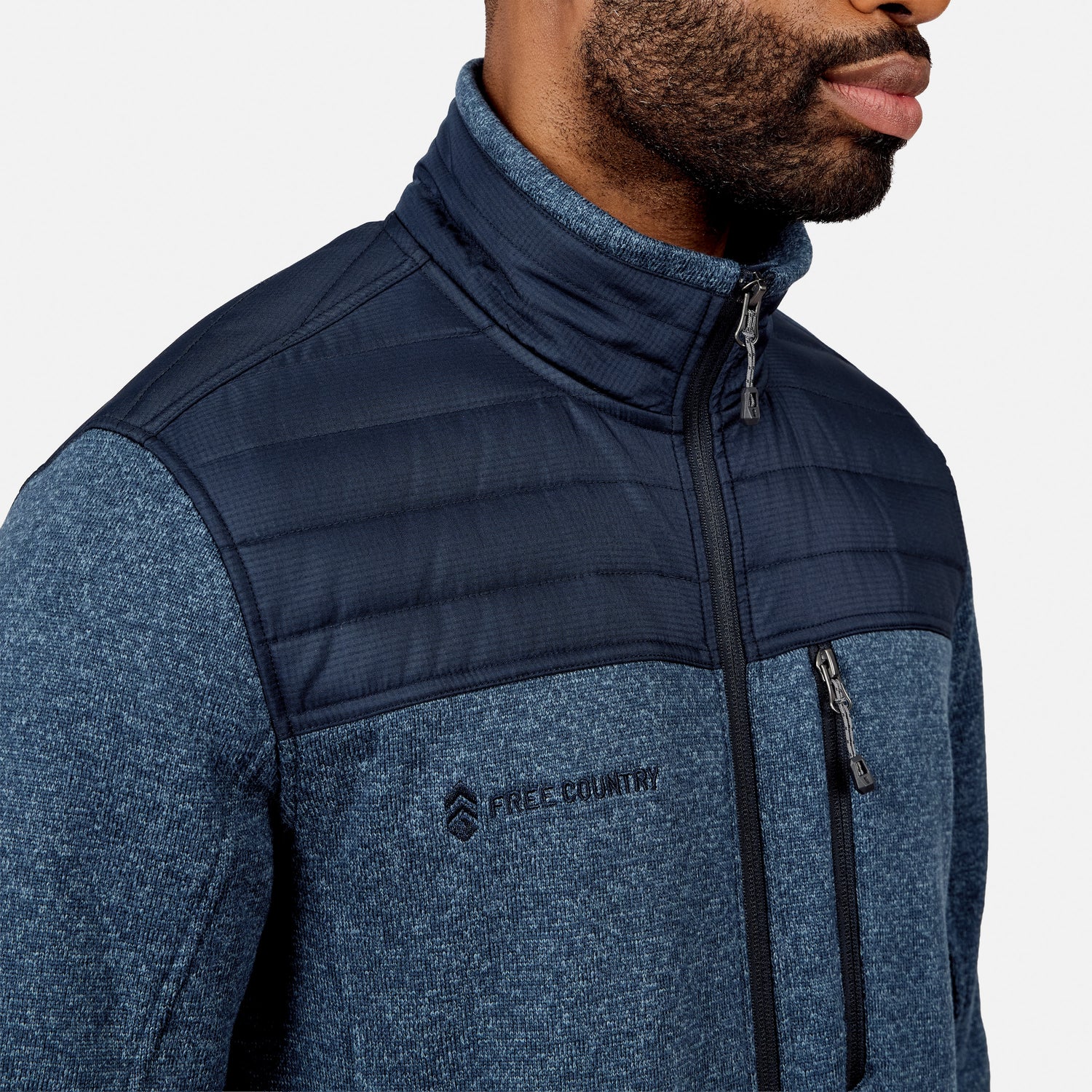 A man wearing the Free Country Men’s Frore Knit Fleece Jacket, featuring a blue knit design, zippered chest pocket, and quilted upper panel, is shown from the shoulders up against a plain background.