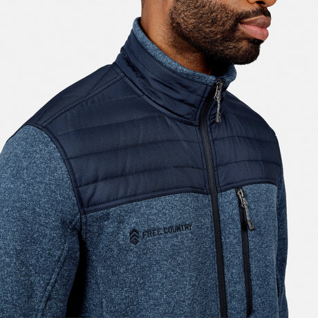 A man wearing the Free Country Men’s Frore Knit Fleece Jacket, featuring a blue knit design, zippered chest pocket, and quilted upper panel, is shown from the shoulders up against a plain background.