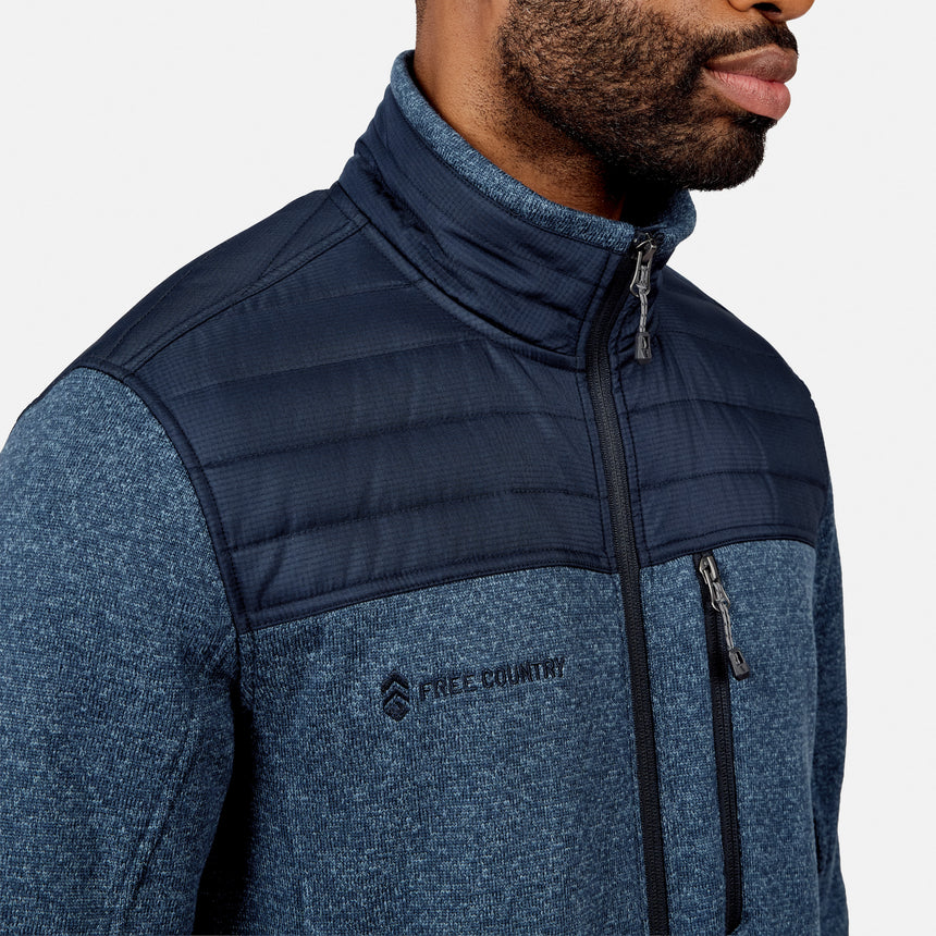 A man wearing the Free Country Men’s Frore Knit Fleece Jacket, featuring a blue knit design, zippered chest pocket, and quilted upper panel, is shown from the shoulders up against a plain background.