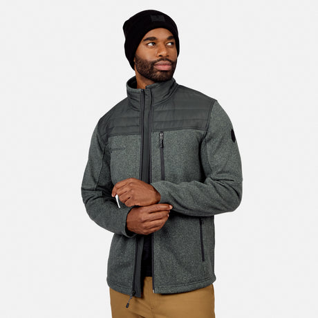 A man in a dark green Free Country Men’s Frore Knit Fleece Jacket, black knit beanie, and tan pants stands against a plain light backdrop, looking left with his hands clasped.