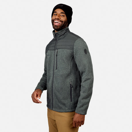 Smiling man in a Free Country Men’s Frore Knit Fleece Jacket (dark gray), black beanie, and tan pants stands against a plain light gray background.