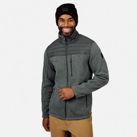A man wears a black beanie, tan pants, and the Free Country Men’s Frore Knit Fleece Jacket—featuring quilted shoulders and a convertible collar—while smiling against a plain white background.
