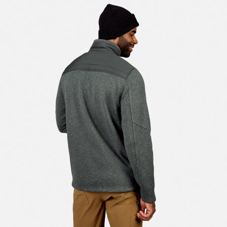 Man in a dark beanie and tan pants models the Free Country Men’s Frore Knit Fleece Jacket, featuring a convertible collar and quilted shoulders, as he faces away and looks back over his right shoulder against a plain light background.