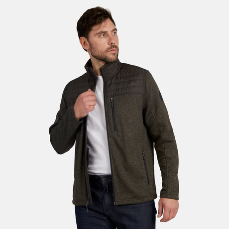 A man wearing the Free Country Men's Frore Knit Fleece Jacket in dark olive, featuring a quilted overlay and convertible collar, pairs it with a white t-shirt and dark jeans as he stands against a plain light background, adjusting his collar.