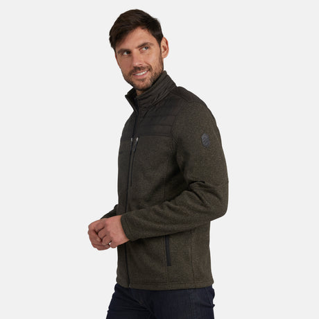 A man with short brown hair and a beard models the Free Country Men's Frore Knit Fleece Jacket, featuring dark green fleece and black quilted shoulders, as he stands sideways against a plain light background.