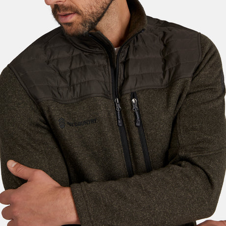 A man wearing the Free Country Men's Frore Knit Fleece Jacket in dark olive green, featuring a convertible collar and quilted shoulder and chest panels, stands with arms crossed, gazing slightly away from the camera.