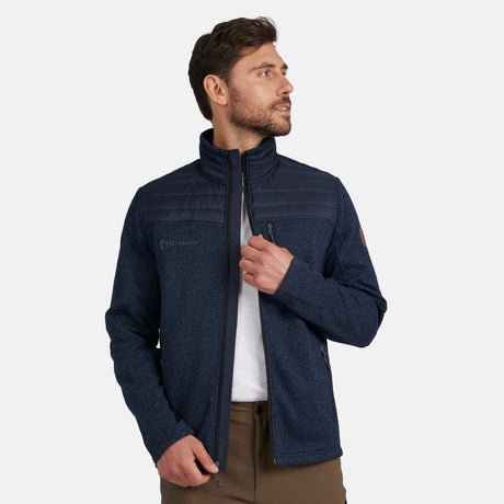 A man with brown hair and a beard, wearing the Free Country Men's Frore Knit Fleece Jacket in navy blue with quilted shoulder panels and a convertible collar, pairs it with brown pants while looking left against a plain light background.