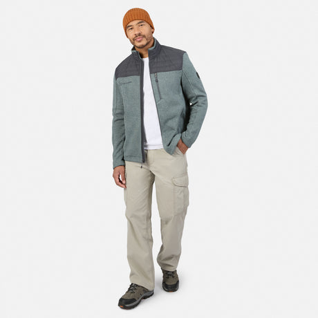 A man wears a grey Men’s Frore Knit Fleece Jacket with a charcoal chest panel and zippered pockets over a white t-shirt, paired with tan cargo pants and a burnt-orange beanie.