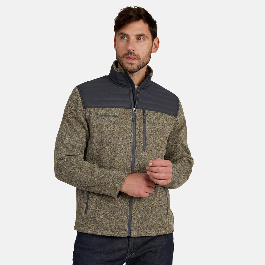 A man wearing the Free Country Men’s Frore Knit Fleece Jacket, featuring a gray and olive colorway, convertible collar, dark shoulder panels, and zippered pockets, poses with hands clasped against a plain white background.
