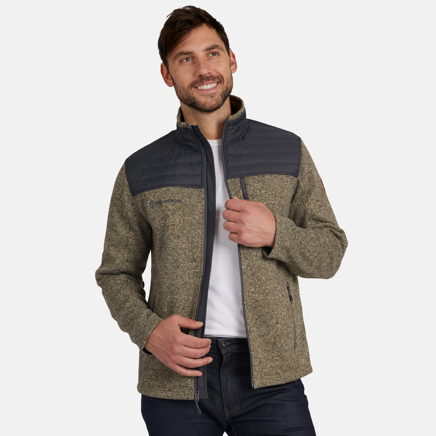 A man with brown hair and a beard stands smiling and facing slightly to the side, zipping up his Men’s Frore Knit Fleece Jacket by Free Country, which features a gray quilted yoke.