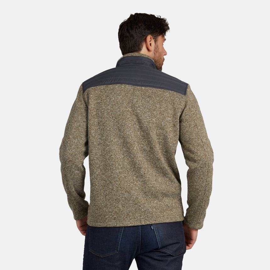 A man, seen from behind, is wearing the Free Country Men’s Frore Knit Fleece Jacket—a textured olive-green jacket with a dark gray shoulder yoke and convertible collar, featuring zippered pockets—paired with blue jeans and set against a plain light background.