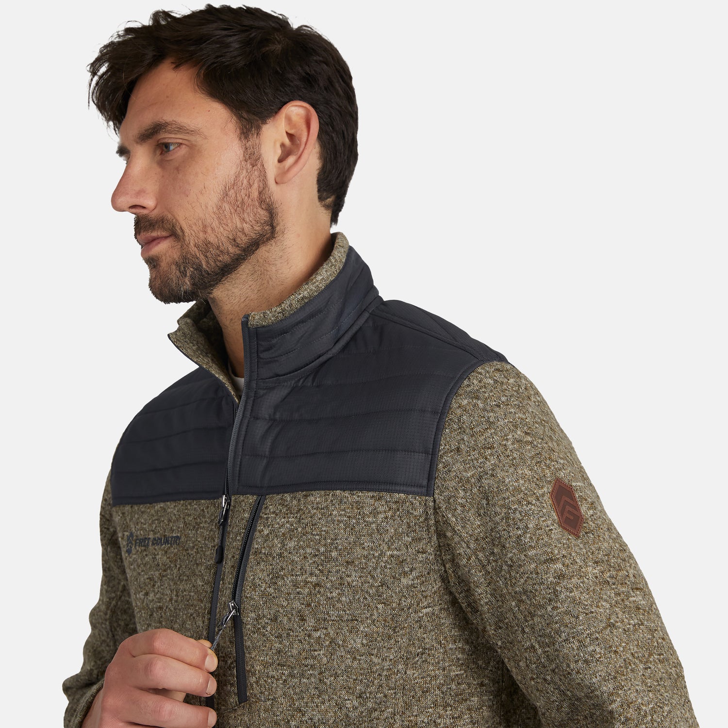 A man with short dark hair and a beard wears the Free Country Men’s Frore Knit Fleece Jacket, featuring quilted shoulders, zippered pockets, and a logo patch on the sleeve. The convertible collar adds versatility as he looks to the side against a light background.