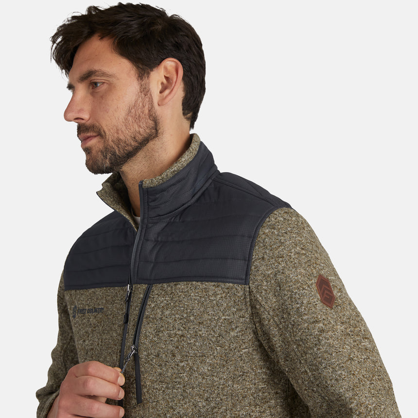 A man with short dark hair and a beard wears the Free Country Men’s Frore Knit Fleece Jacket, featuring quilted shoulders, zippered pockets, and a logo patch on the sleeve. The convertible collar adds versatility as he looks to the side against a light background.